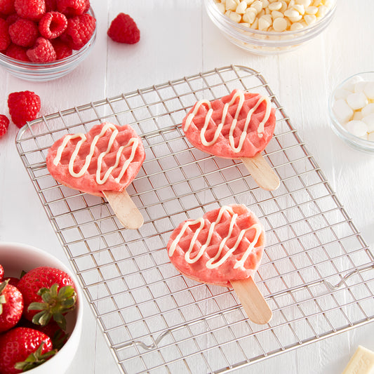 Three pink mini heart waffles with white frosting on popsicle sticks sit on a cooling rack.