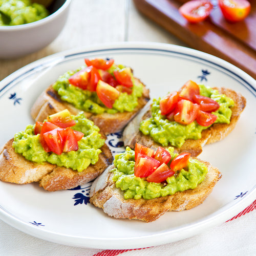 Four pieces of toast with avocado and tomato on top on a white plate.