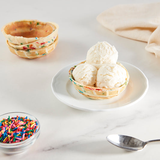 A rainbow sprinkle waffle bowl filled with three scoops of vanilla ice cream on a white plate.