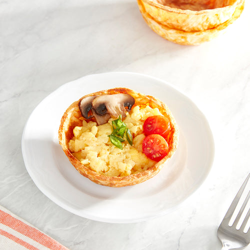 A hashbrown waffle bowl filled with scrambled eggs, tomato, and mushroom, garnished with chives, on a white plate.