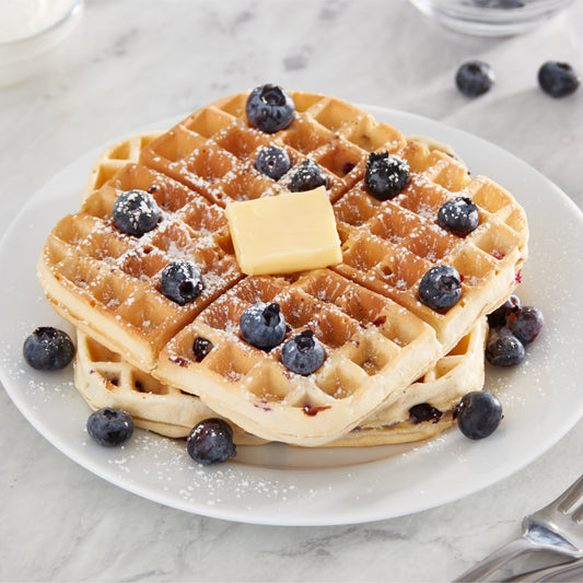 Two 7" waffles on a white plate topped with blueberries, powdered sugar, and a slab of butter.