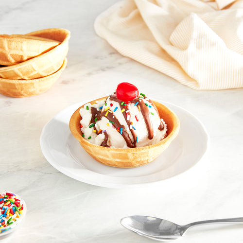 A waffle bowl filled with one scoop of vanilla ice cream, chocolate drizzle, rainbow sprinkles, and a cherry on top on a white plate.