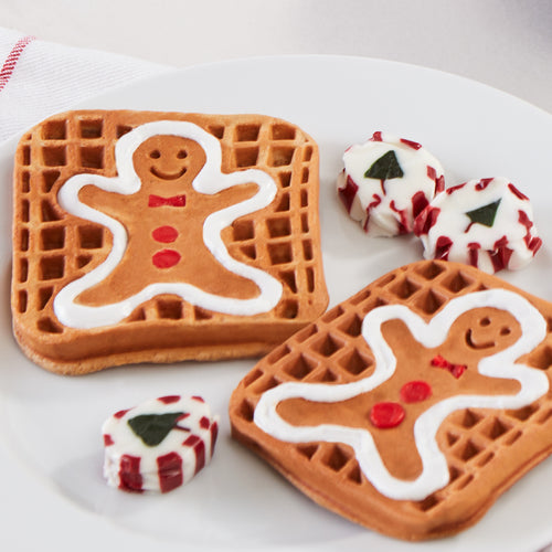 Two waffles with gingerbread man prints with white frosting on a white plate with peppermint candies.