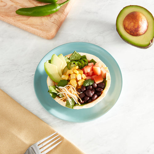 A taco bowl filled with lettuce, tomato, cheese, corn, avocado, jalapenos, corn, and black beans, on a blue plate.