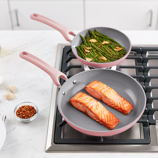 Two frying pans on a stove, one has two salmon fillets and the other has asparagus.