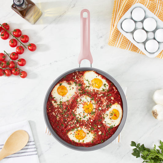 A pan of shakshuka on a white marble countertop next to eggs and tomatoes.