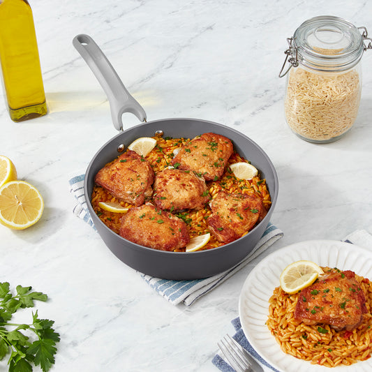 Chicken thighs and orzo in a 3qt sauté pan on a marble countertop.