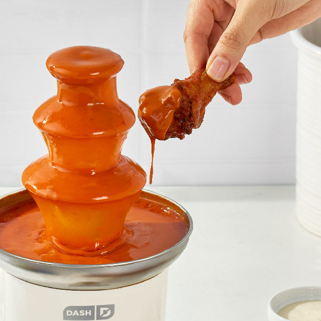 Person dipping a piece of chicken into an orange sauce fountain with 'DASH' branding.
