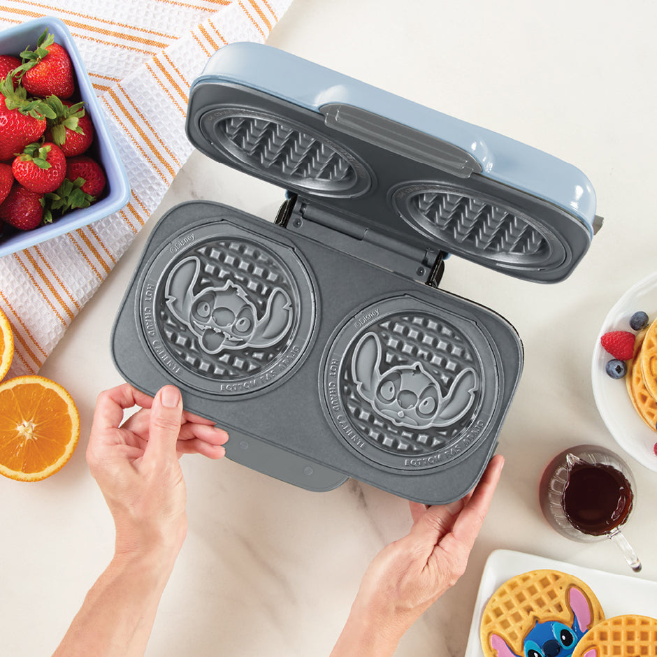 Gray waffle maker with Stitch character designs on a table with waffles and fruit.