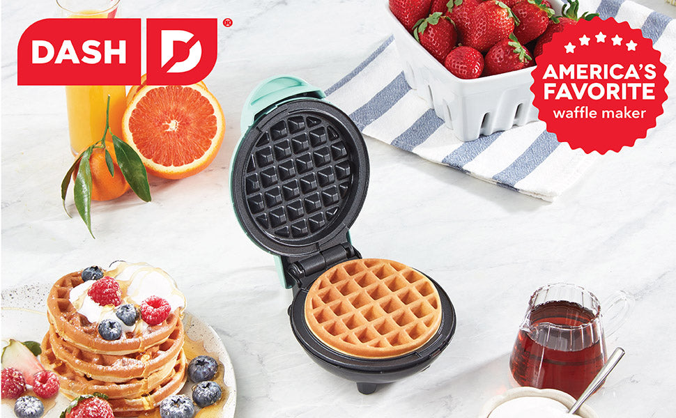A mint green Dash mini waffle maker sits open with a golden waffle inside. Fresh fruit, syrup, and waffles surround it on a marble surface. A red badge reads “America’s Favorite Waffle Maker"