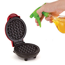 Waffle maker with a hand holding a spray bottle with yellow liquid on a white background