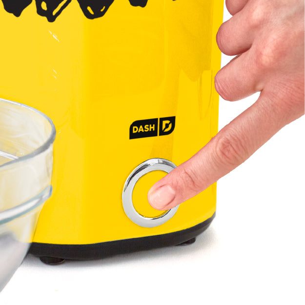 Finger pressing power button on yellow Peanuts® x Dash Popcorn Maker to start popping.