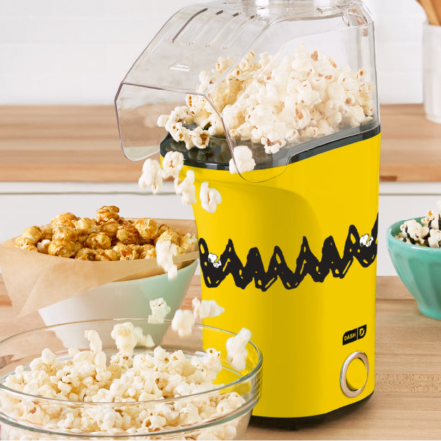 Peanuts® x Dash Popcorn Maker in yellow Charlie Brown design popping popcorn on kitchen counter.