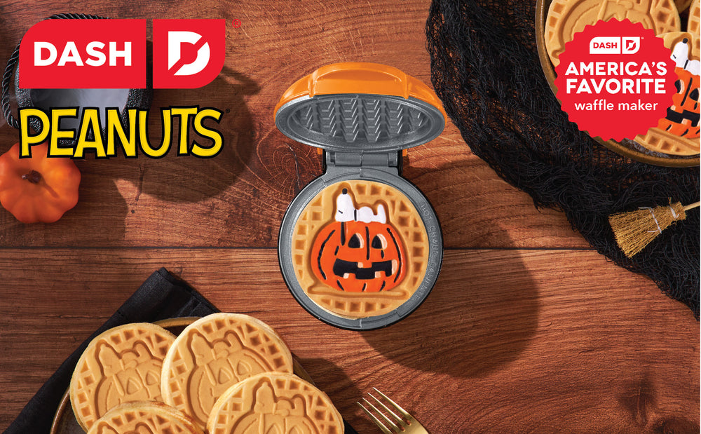 Waffle maker with Halloween-themed waffle on a wooden surface, surrounded by pumpkins and broomsticks.