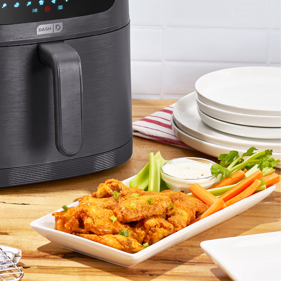 Black Dash air fryer on a wooden table with a plate of fried chicken, carrots, celery, and dip.