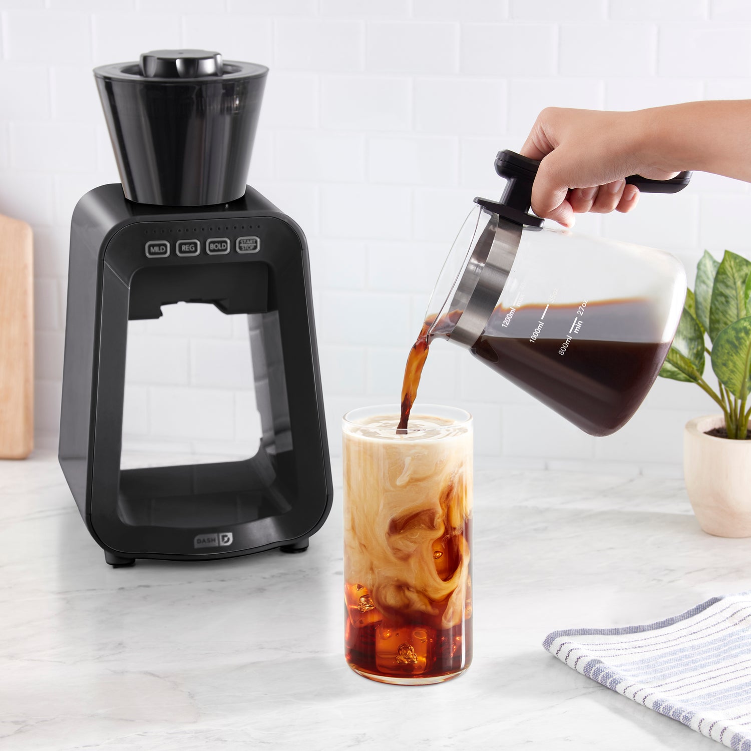 A pitcher of cold brew coffee being poured into a tall clear glass with milk.