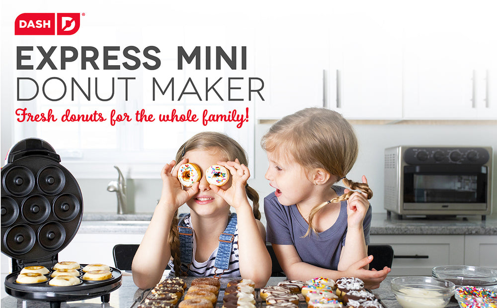 2 young girls standing at a counter full of mini donuts. One twirls her hair braid and looks at the other who is holding donuts up to her eyes as glasses.