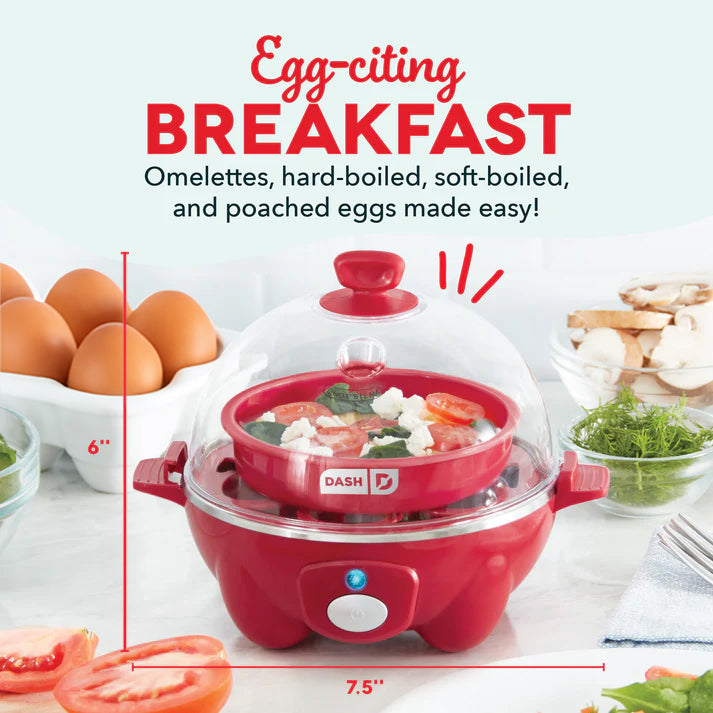 Red and white Dash egg cooker with clear dome lid, surrounded by eggs, tomatoes, and fresh ingredients on white surface.