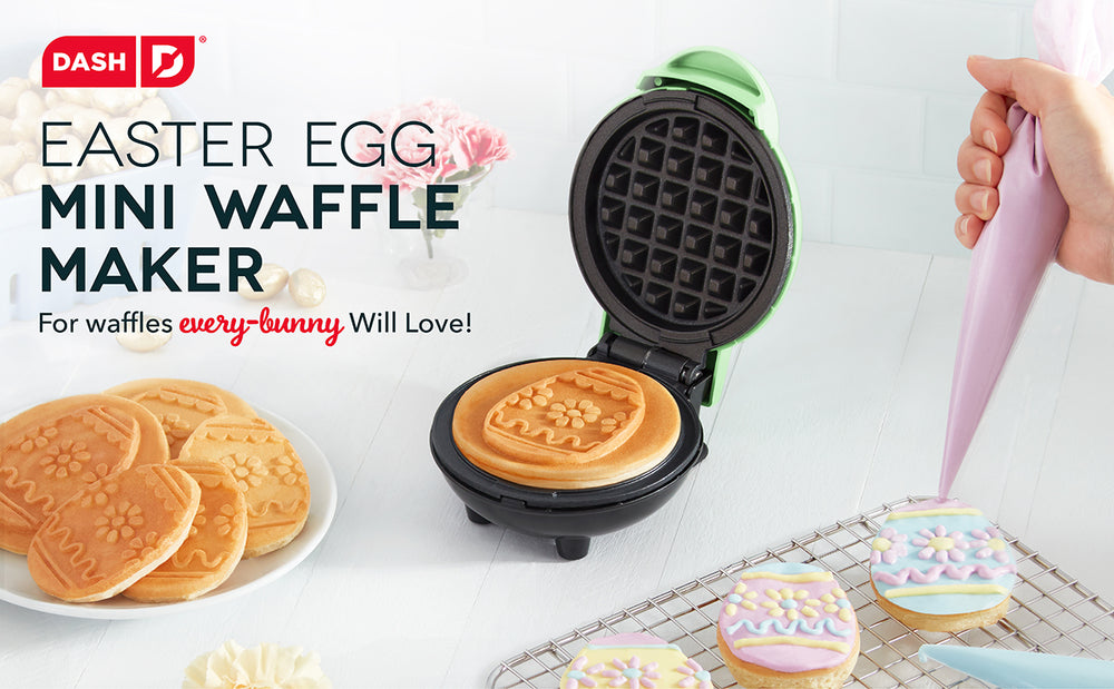 A mint colored Easter Egg Mini Waffle Maker sits open with a fresh waffle alongside a cooling rack with frosted waffles.