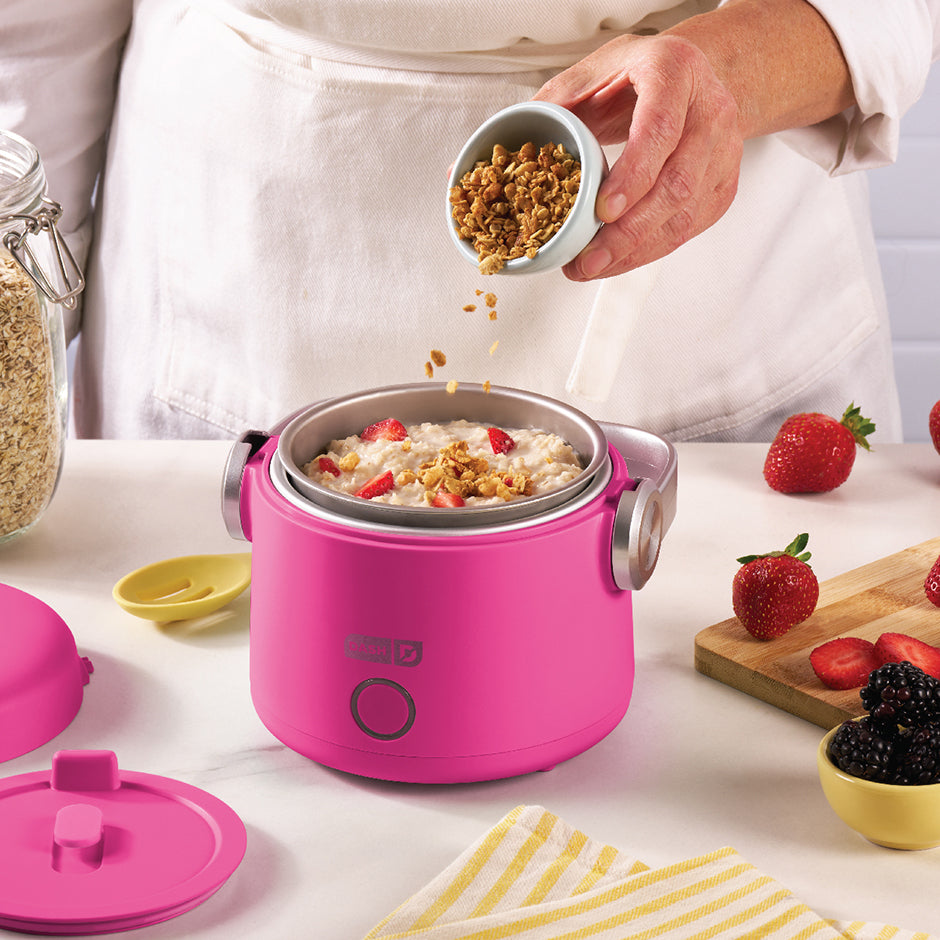 Granola being sprinkled over oatmeal with strawberries inside the pink Dash Hot2Go™ Electric Lunchbox—perfect for a warm, homemade breakfast.
