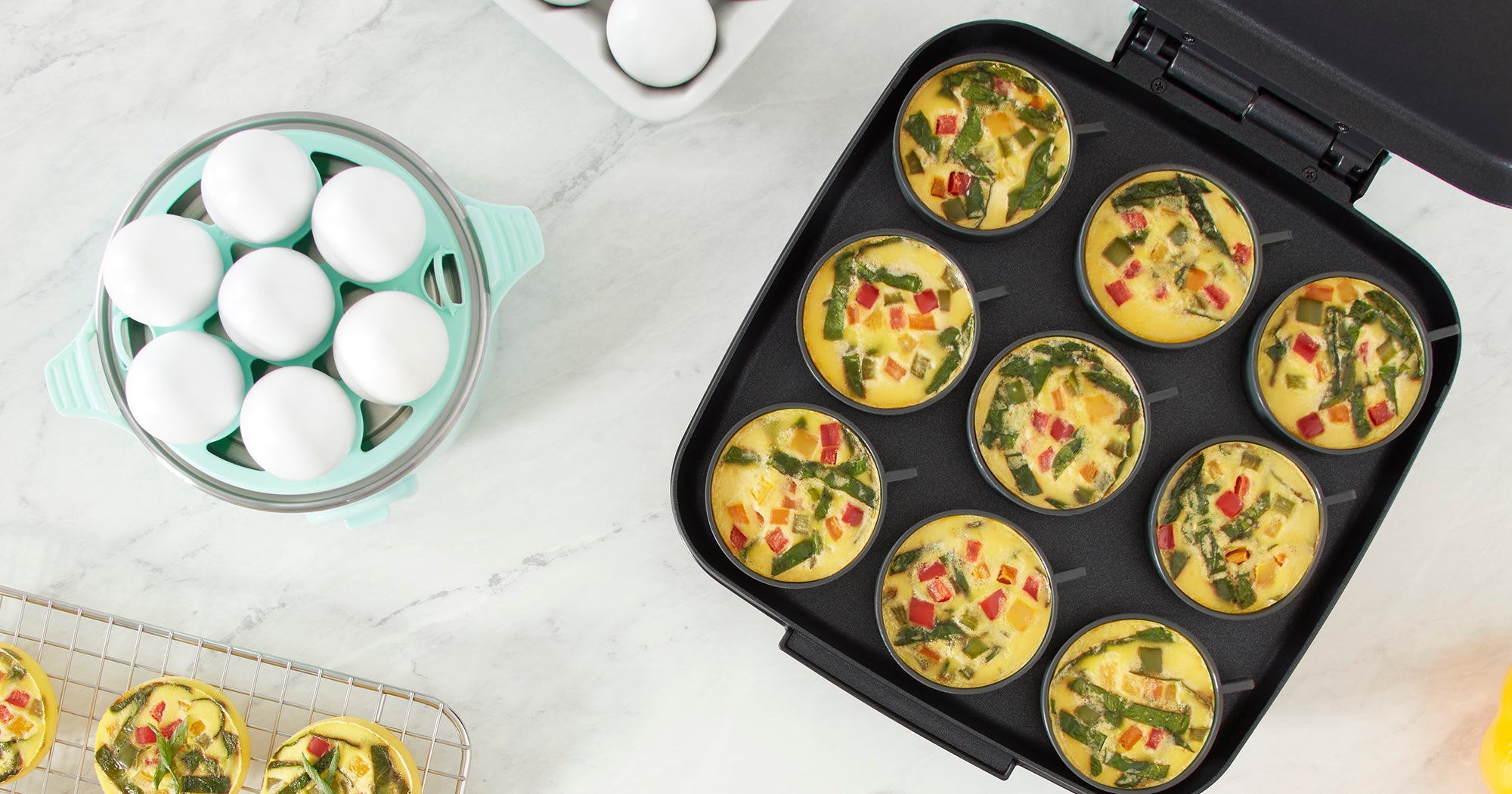 The Dash Rapid Egg Cooker and Family Size Egg Bite Maker on a marble countertop with veggie egg bites and hard boiled eggs.