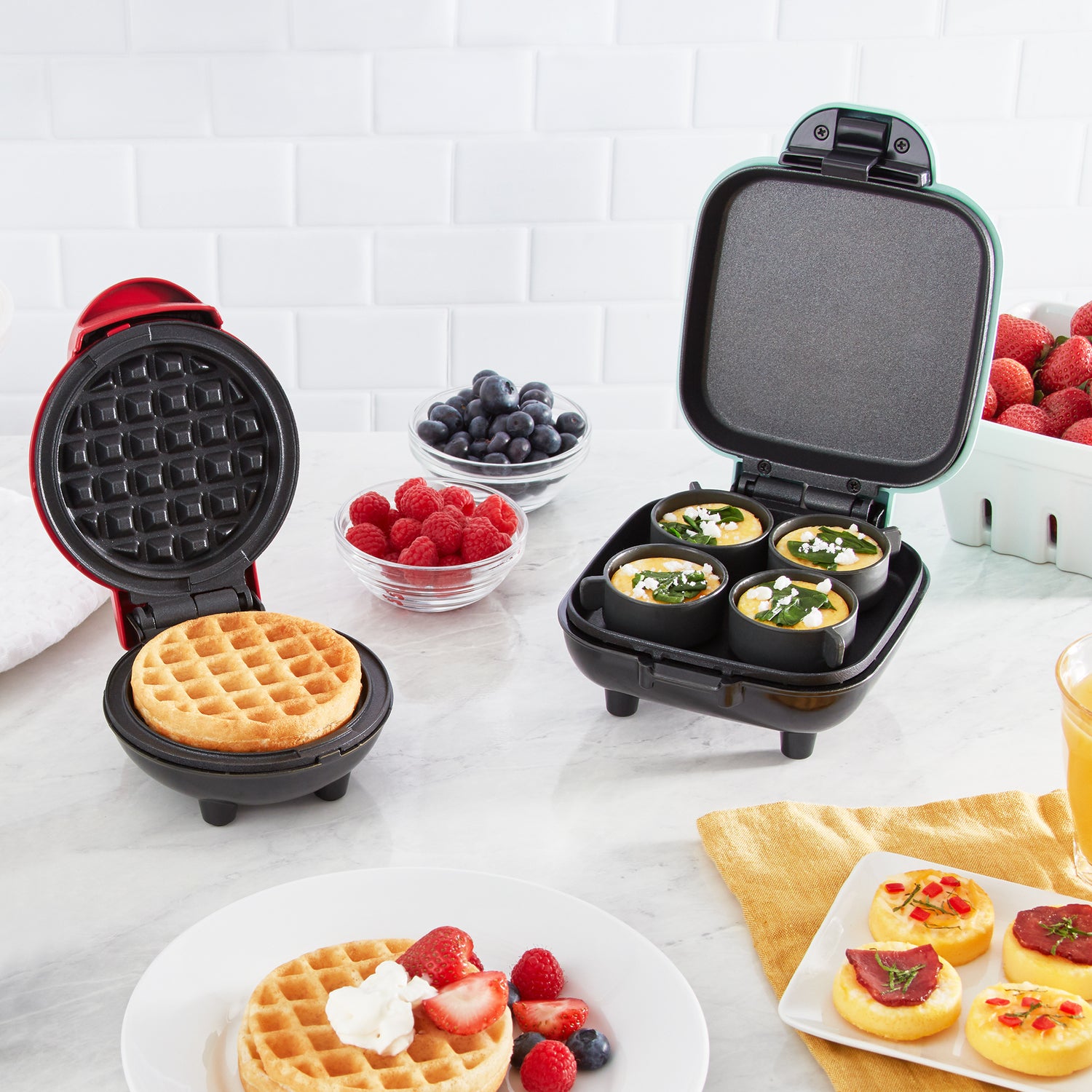 A red Dash Mini Waffle Maker and aqua Egg Bite Maker on a white counter.