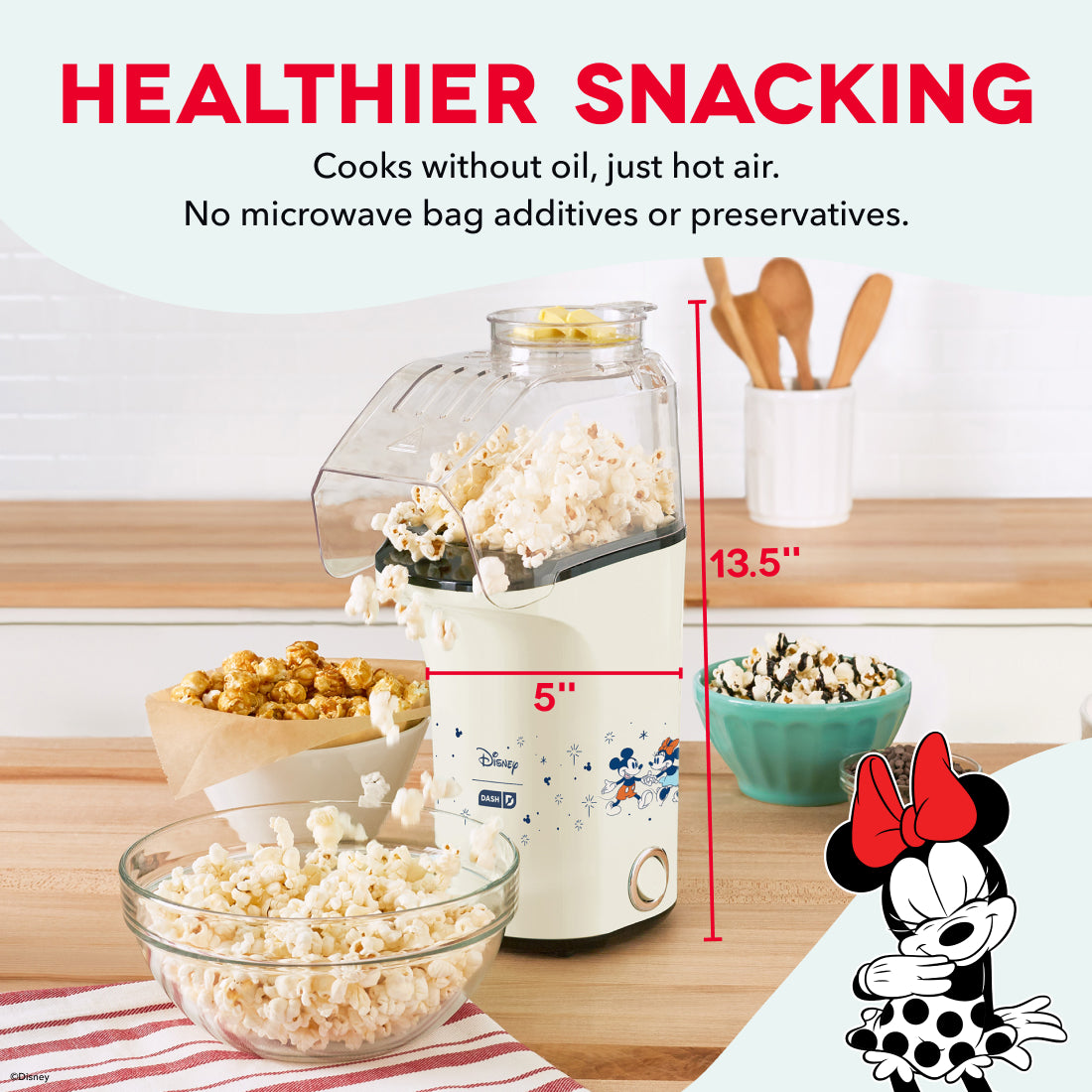 Cream-colored popcorn maker with clear top chamber, surrounded by bowls and containers of popped popcorn on wooden surface.