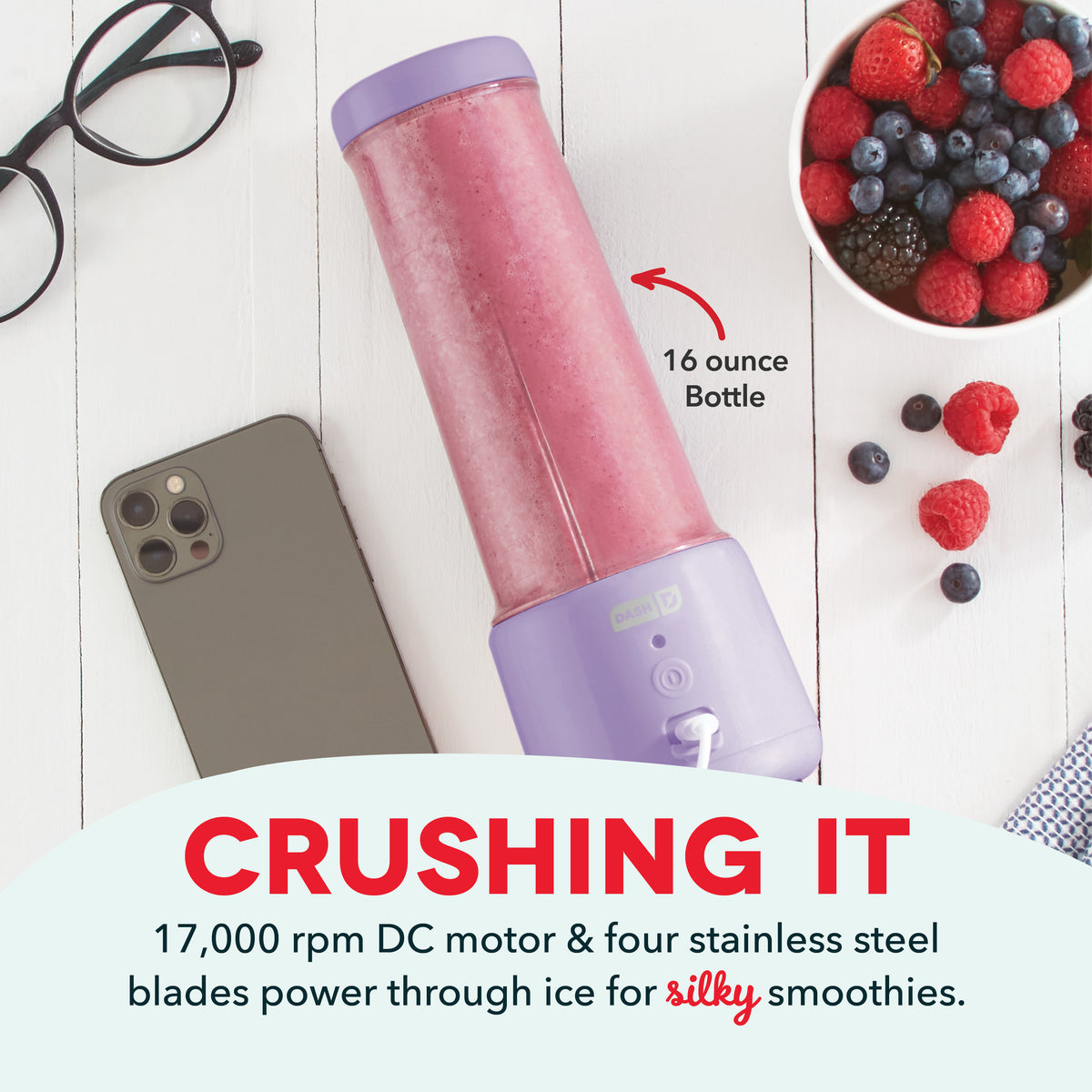 Pink and purple 16-ounce portable blender with purple lid next to smartphone, berries, and glasses on white wooden surface.